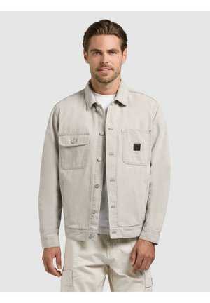 Mann trägt eine hellbeige, geknöpfte Jacke mit zwei Brusttaschen und passende Cargo-Hosen, stehend vor einfarbigem Hintergrund.