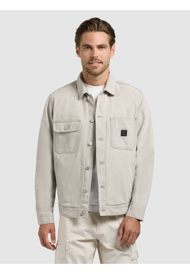Homme portant une veste boutonnée beige clair avec deux poches poitrine et un pantalon cargo assorti, debout devant un fond uni.