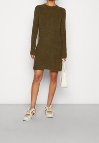 Abito lavorato a maglia verde oliva con maniche lunghe e scollatura rotonda, indossato con sneaker beige e bianche. Modella che tiene una borsa bianca.