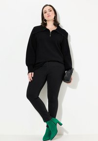 Suéter negro con cuello con cremallera, textura acanalada, combinado con jeans ajustados negros. Botines de ante verdes hasta el tobillo y un pequeño clutch negro.