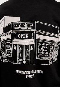 Sort sweatshirt med et hvidt grafisk motiv af en butik og teksten "DEF", "OPEN" og "WORKATION COLLECTION F/W23."