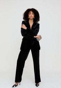 Schwarzer samtiger Blazer und Hose mit weitem Bein, mit tiefem V-Ausschnitt, kombiniert mit gepunkteten High Heels und einer gestreiften Clutch.