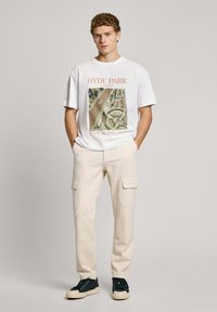 Witte t-shirt met druk "HYDE PARK LONDEN" en kleurrijke illustratie. Beige cargo shorts met zijzakken. Zwarte sneakers met een witte zool.