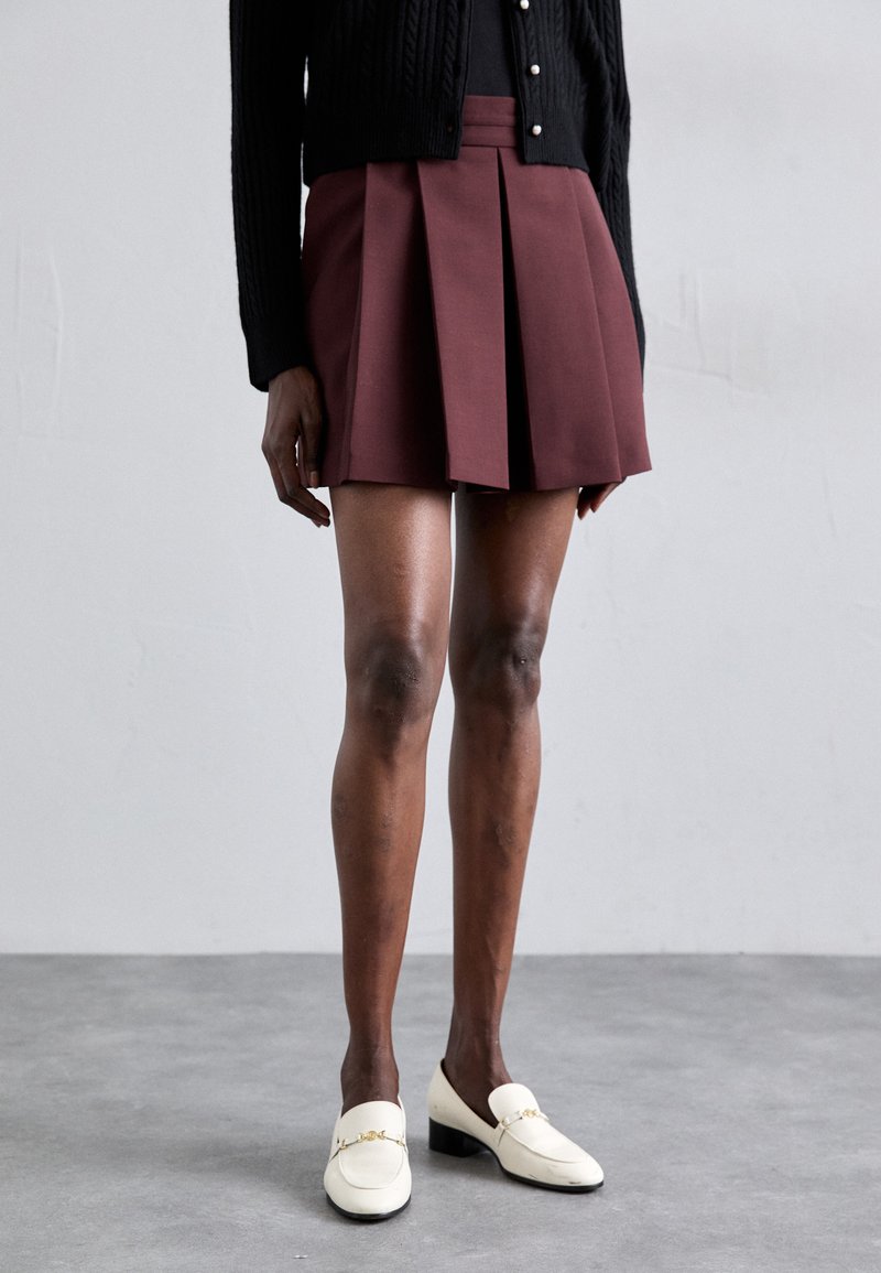 Jupe plissée bordeaux associée à un cardigan noir. Des mocassins blancs avec des accents dorés complètent la tenue.