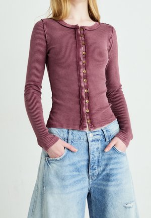 Frau trägt ein eng anliegendes, geripptes, mauvefarbenes Langarmshirt mit Knöpfen, kombiniert mit locker sitzenden, hellblauen Jeans mit Händen in den Taschen.