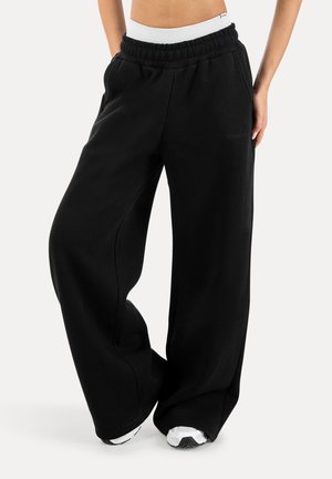 Losse zwarte joggingbroek met elastische tailleband en zijzakken, gedragen met witte sneakers, onderlichaam en benen zichtbaar tegen een witte achtergrond.