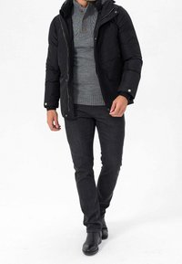 Sort polstret jakke med hætte, grå striktrøje med høj krave, mørke jeans og sorte støvler. Bemærkelsesværdige knapdetaljer på trøjen.