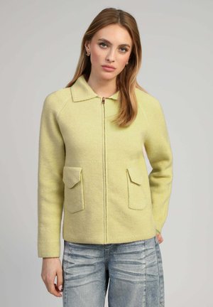 Jeune femme aux cheveux châtain clair portant une veste zippée jaune pâle avec deux poches frontales et un jean délavé bleu clair.