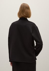 Persona con i capelli castano chiaro corti, che indossa una maglietta nera a maniche lunghe larga, in piedi con la schiena rivolta verso la fotocamera davanti a un muro chiaro e semplice.