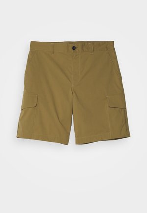 Mies, jolla on musta poolopaita, oliivinvihreät cargo-shortsit, mustat sukat ja mustat platvormikengät, seisoo harmaalla lattialla vaaleanharmaata seinää vasten.