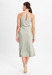 Satin grüne Midi-Kleid mit einem Schlüsselaugenrücken, dünnen Trägern und einem ausgestellten Saum. Getragen mit silbernen High Heels, die ein glattes Finish zeigen.