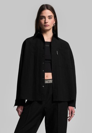 Camicia nera oversize realizzata in tessuto testurizzato, con chiusura con bottoni, colletto e tasca sul petto sinistro. Indossata sopra un crop top nero.