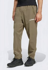 Pantalons d'extérieur khaki avec une coupe décontractée, taille élastique, poches zippées sur les côtés et logo blanc "TERREX" sur la cuisse gauche. Portés avec des chaussures noires.