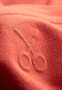 Tissu corail avec une surface texturée arborant un motif de ciseaux brodés. La broderie est subtile et se fond dans le matériau.