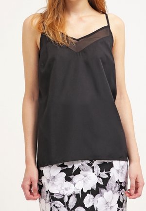 Woman wearing a black sleeveless sheer V-neck top paired with a black and white floral skirt, standing against a plain background.