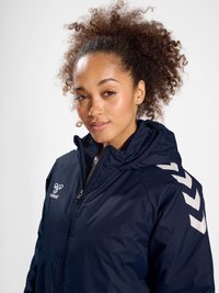 Vrouw met krullend haar draagt een navy blauwe jas met rits en capuchon en witte Chevron-logo's op de schouder en borst.