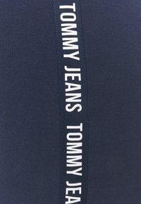 Mörkblått tyg med en vertikal vit rand och texten "TOMMY JEANS". Materialet har en slät yta och en kroppsnära design.