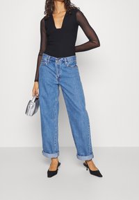 Zwarte bodysuit met doorschijnende lange mouwen, hoge halslijn, gecombineerd met blauwe, loszittende jeans met opgerolde mouwen en zwarte puntige hakken. Zilveren tas.
