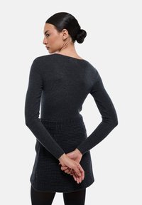 Pull en maille gris foncé à manches longues avec un large décolleté, associé à une jupe noire texturée, vu de dos avec les mains croisées.