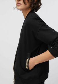 Must bomberjakk lukuga, millel on soonikkoes varrukasäärmed ja kuldsete ketti detailidega tasku. Sile materjal ja mugav lõige.