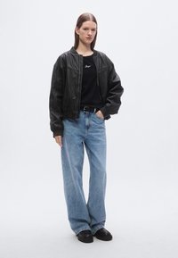 Blouson aviateur en cuir noir avec poignets côtelés, associé à un T-shirt noir et un jean baggy taille haute bleu clair. Chaussures noires épaisses.