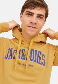 Gelbe Kapuzenjacke mit Kordelzug, mit blauer Aufschrift "JACK & JONES intl. seit 1990." Glatte Textur mit gerippten Bündchen.