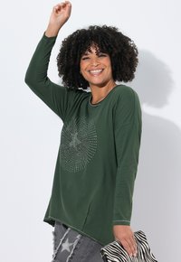 Chemise verte à manches longues avec un col rond et un motif décoratif à l'avant en points métalliques, associée à un jean gris déchiré et à un sac rayé.