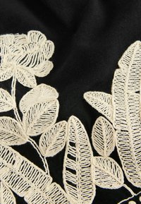 Broderie florale couleur crème avec des feuilles sur un tissu noir, montrant des motifs de fil détaillés et une texture.