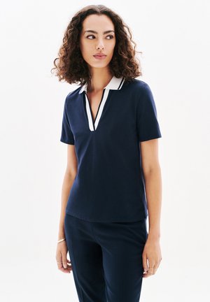 Polo shirt blu navy con un bordo bianco al colletto e un leggero scollo a V. Maniche corte, texture liscia, design aderente, stile minimalista.