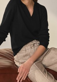 Pullover a coste nera con colletto polo, abbinato a pantaloni beige chiaro a vita alta con texture, appoggiato su una superficie in pelle marrone.