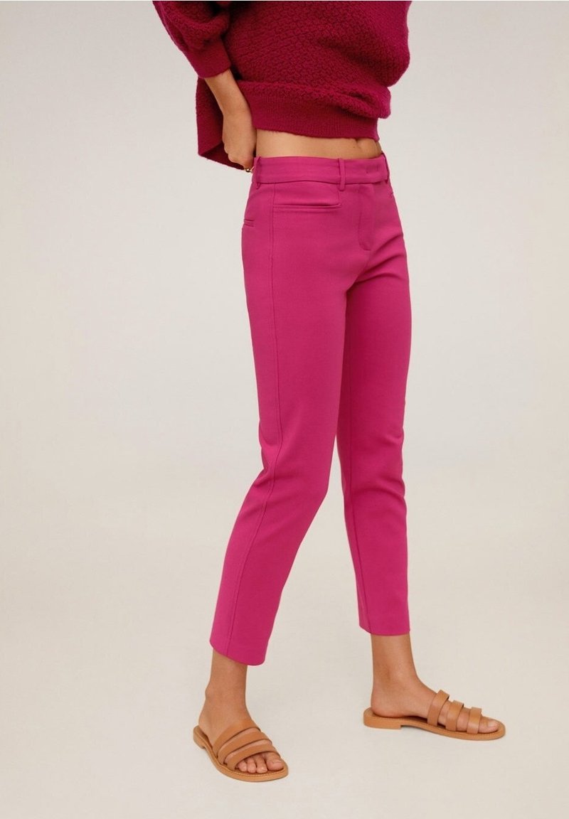Femme portant un pantalon rose vif jusqu'aux chevilles, des sandales plates couleur camel, et un pull texturé bordeaux, debout devant un fond uni.