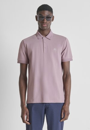 Joven con gafas de sol cuadradas y oscuras, una camiseta tipo polo malva con logo y pantalones azul marino, posando frente a un fondo liso.