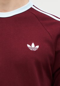 Μπορντό φούτερ με λευκό γιακά και ρίγες στους ώμους, με μικρό λευκό λογότυπο τριφυλλιού στο στήθος.