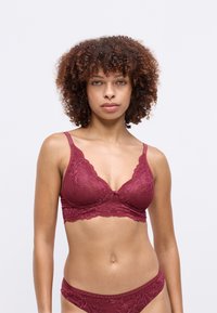 Donna con capelli ricci che indossa un reggiseno e una mutandina in pizzo rosso scuro abbinati, rivolta in avanti su uno sfondo chiaro e semplice.