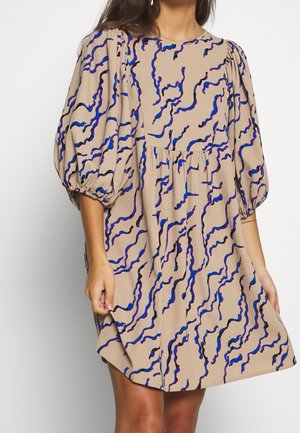 Kobieta w beżowej sukience z niebiesko-czarnym abstrakcyjnym falistym wzorem, z bufiastymi rękawami długości do łokcia i marszczonym detalem w talii.