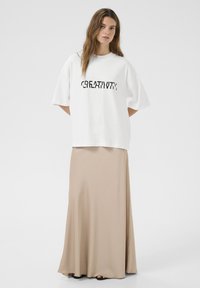 Witte oversized t-shirt met zwarte tekst "CREATIVTY," gecombineerd met een lange, beige, losse rok. Glad materiaal en eenvoudig ontwerp.