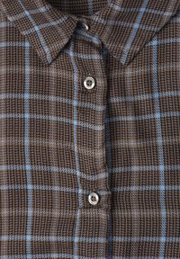 Gros plan sur une chemise à carreaux marron et bleue avec un tissu texturé et deux boutons noirs et argentés visibles sur la patte de devant.