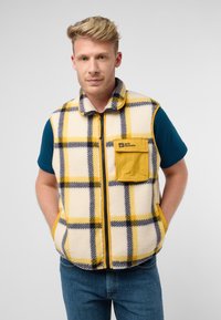 Gilet in pile con motivo a quadri giallo, nero e crema; presenta una chiusura con zip, colletto alto e tasca applicata. Indossato sopra una camicia blu.