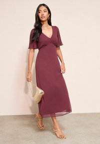 Fialová, stredne dlhá šaty s hlbokým výstrihom do V, flutter rukávmi a detailmi na gombíky. Nosené so zlatými sandálmi na podpätku a metalickou kabelkou.
