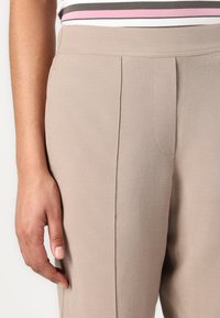 Světlé taupe kalhoty s hladkou texturou, s předními sklady a přizpůsobeným střihem. Pás je elegantní a sedí nad boky.