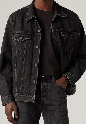 Giacca di denim nera con due tasche sul petto, chiusure con bottoni argentati e cuciture a contrasto. Indossata sopra una t-shirt nera e jeans.