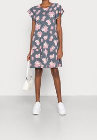 Blomstermönstrad grå klänning med korta volangärmar, lager-på-lager-design och mjukt tyg. Kombineras med vita sneakers och en liten vit handväska.