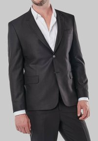 Veste de costume noire ajustée avec une texture lisse, fermeture à un bouton et revers crantés, associée à une chemise habillée blanche en dessous.