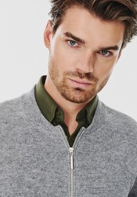 Szary sweter z dzianiny z zamkiem z przodu, noszony na ciemnozielonej koszuli z guzikami. Miękka faktura, dopasowany krój i prążkowany kołnierz.