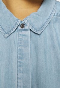 Camicia in denim azzurro chiaro con colletto a punta e un solo bottone nero vicino al collo, con dettagli delle cuciture visibili e tessuto testurizzato.