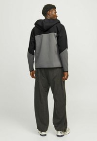 Giacca grigia e nera con cappuccio, caratterizzata da un design a blocchi di colore e una texture liscia. Abbinata a pantaloni scuri di taglio comodo e sneakers.