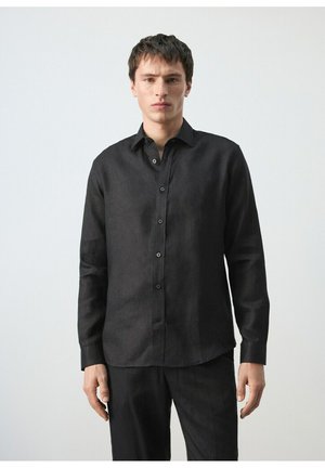 Joven de pie vistiendo una camisa negra de manga larga con botones y pantalones negros a juego contra un fondo claro y sencillo.