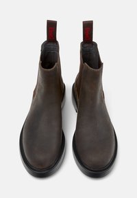Stivaletti alla caviglia in pelle marrone con pannelli elastici laterali, dotati di suola in gomma nera e linguette rosse con il logo sul retro. Texture liscia, punta arrotondata.