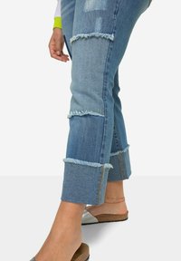 Jeans de denim azul desgastados con dobladillo deshilachado, que presentan parches desgastados verticales. Combinados con unas sandalias de tiras marrones.