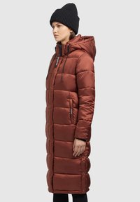 Long, quilted puffer coat in rust color with a hood, front zipper, adjustable drawstrings, and side pockets. Worn with a black knit beanie.